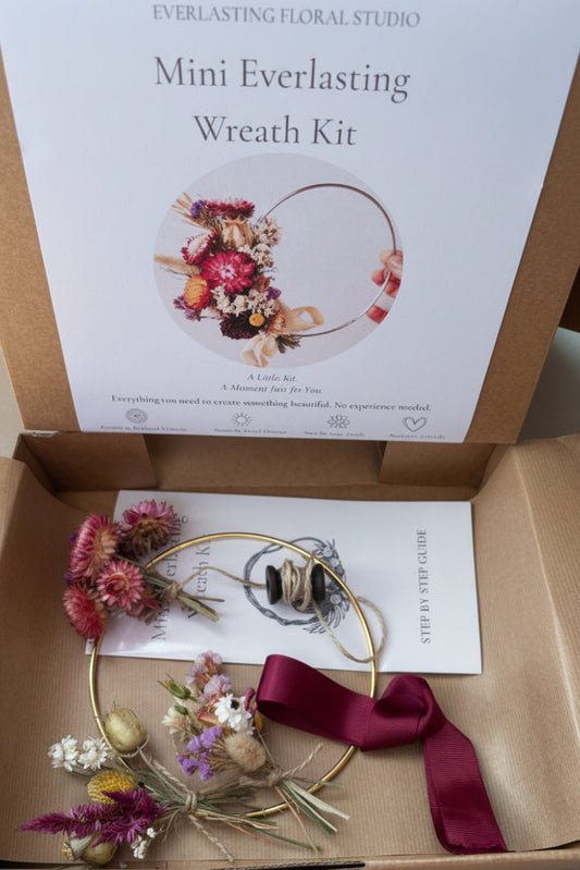 Open cardboard gift box containing a Mini Everlasting Wreath Kit from Everlasting Floral Studio, with a gold wire wreath, dried flowers, a burgundy ribbon, and a printed instruction card.
