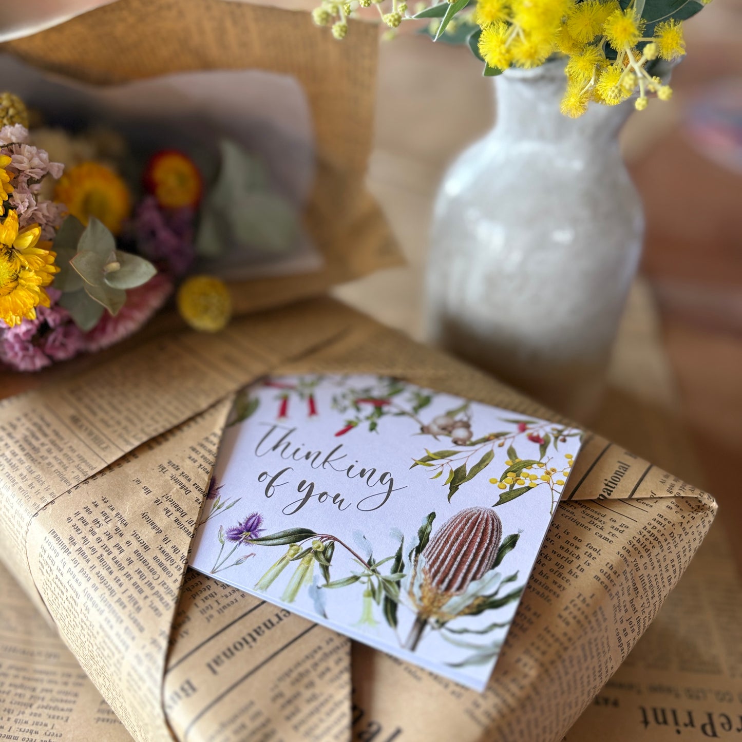 A floral greeting card reading 'thinking of you' with botanical illustrations, placed on top of newspaper-wrapped packaging. A white vase with yellow flowers and another bouquet are softly blurred in the background.