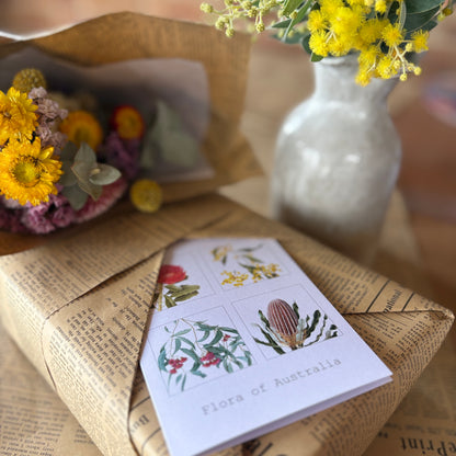 A folded white card titled "Flora of Australia" with botanical illustrations lies on top of a newspaper-wrapped package. In the softly blurred background, yellow flowers in a white vase and another bouquet are visible.