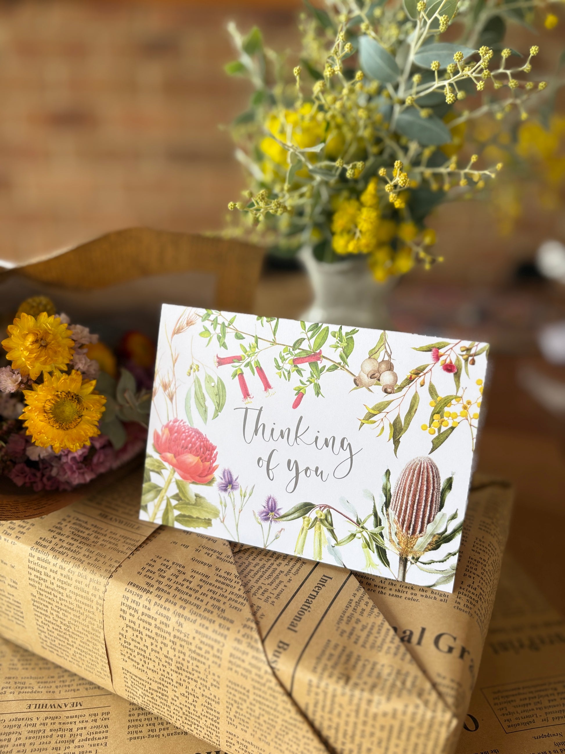 A floral greeting card that reads 'Thinking of you' in elegant script, styled on top of newspaper-wrapped packaging with dried flowers and a blurred vase of yellow flowers in the background.