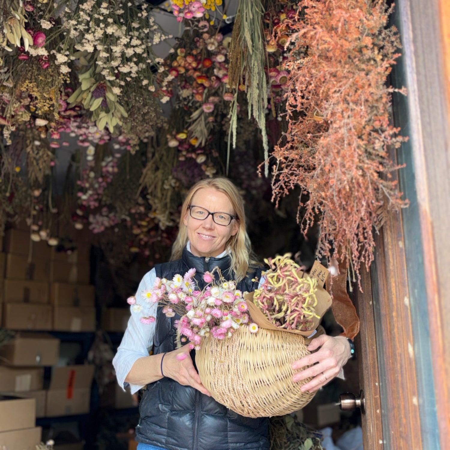 Person holding a basket of flowers in front of a floral shop with dried flowers hanging above.