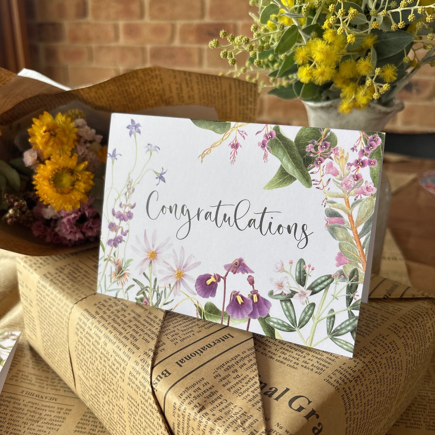 A white congratulations card with watercolor floral illustrations and the word 'Congratulations' in script, styled on top of newspaper-wrapped gift boxes with flowers in the background.