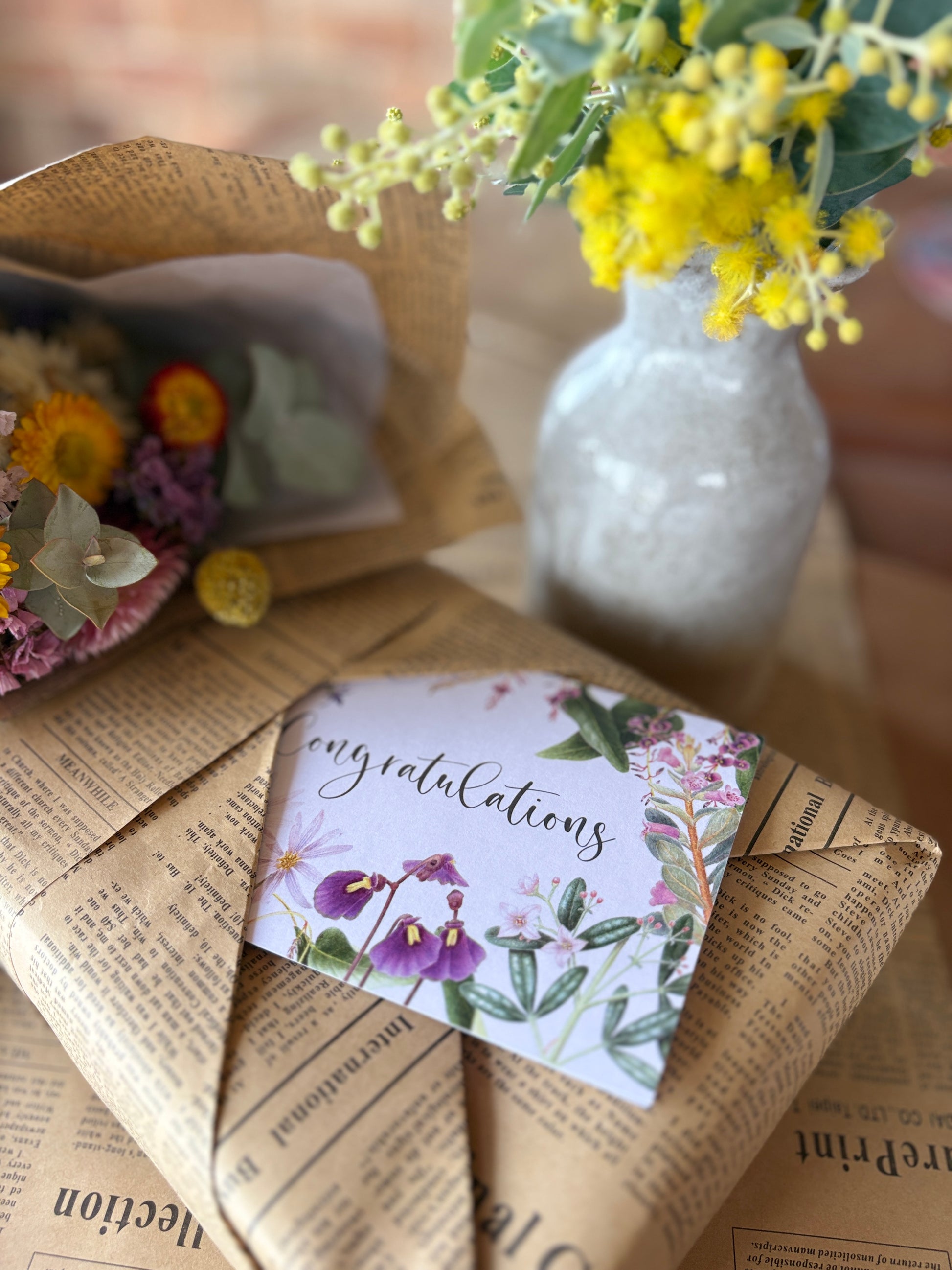 A close-up of a wrapped gift box wrapped in vintage-style brown newspaper print paper, with a floral congratulations card placed on top. A small white vase with yellow flowers sits in the background, and part of another wrapped bouquet is visible on the left.