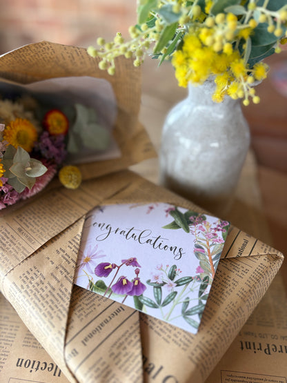 A close-up of a wrapped gift box wrapped in vintage-style brown newspaper print paper, with a floral congratulations card placed on top. A small white vase with yellow flowers sits in the background, and part of another wrapped bouquet is visible on the left.