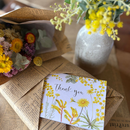 A floral thank you card with yellow flowers and green leaves lies on top of vintage-style newspaper envelopes, with dried flowers and a vase of yellow blossoms in the background.