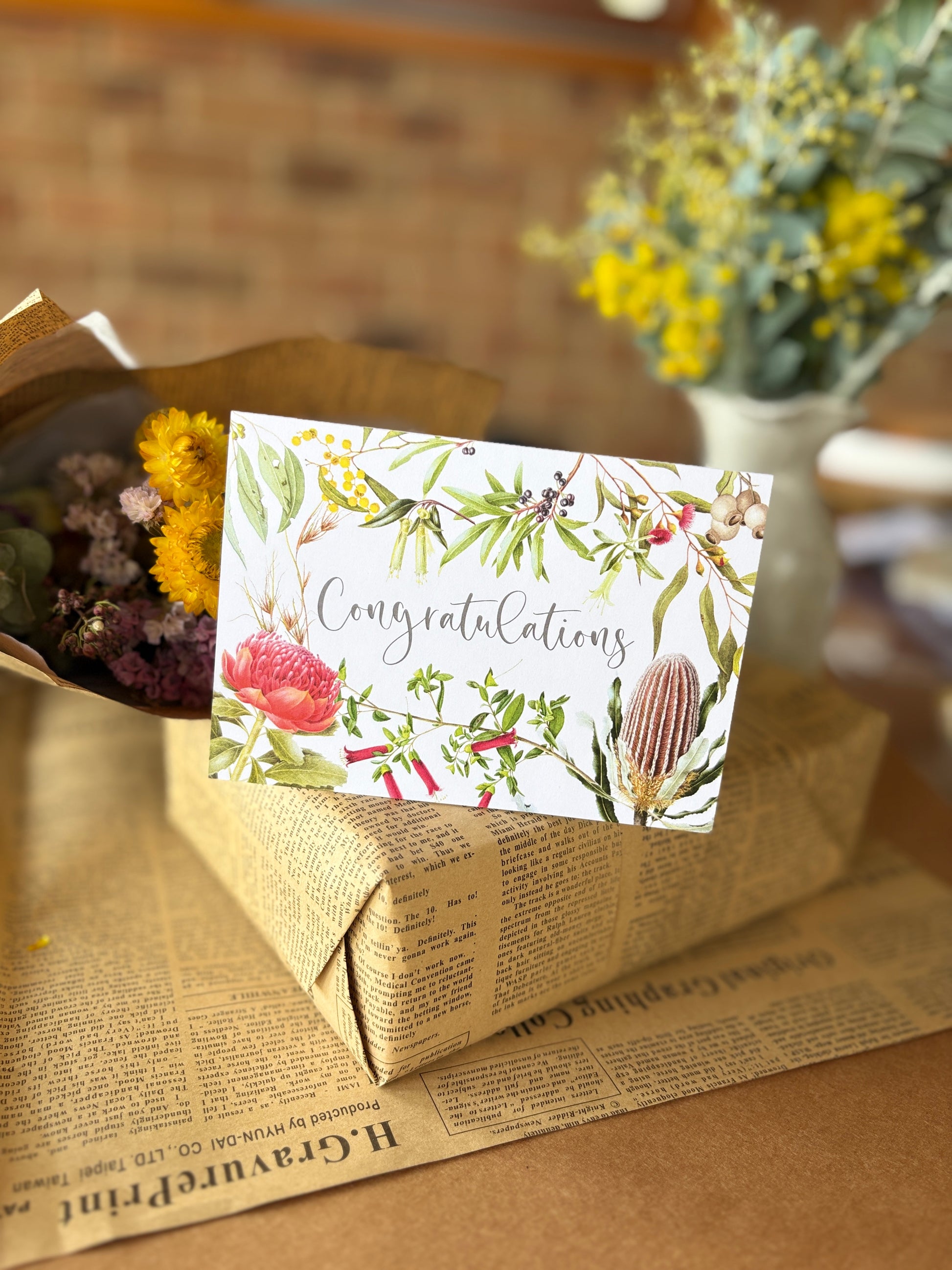A rectangular congratulations card with botanical illustrations and cursive text, resting on a kraft paper gift box wrapped in newspaper-style paper, with dried flowers and a blurred vase in the background.