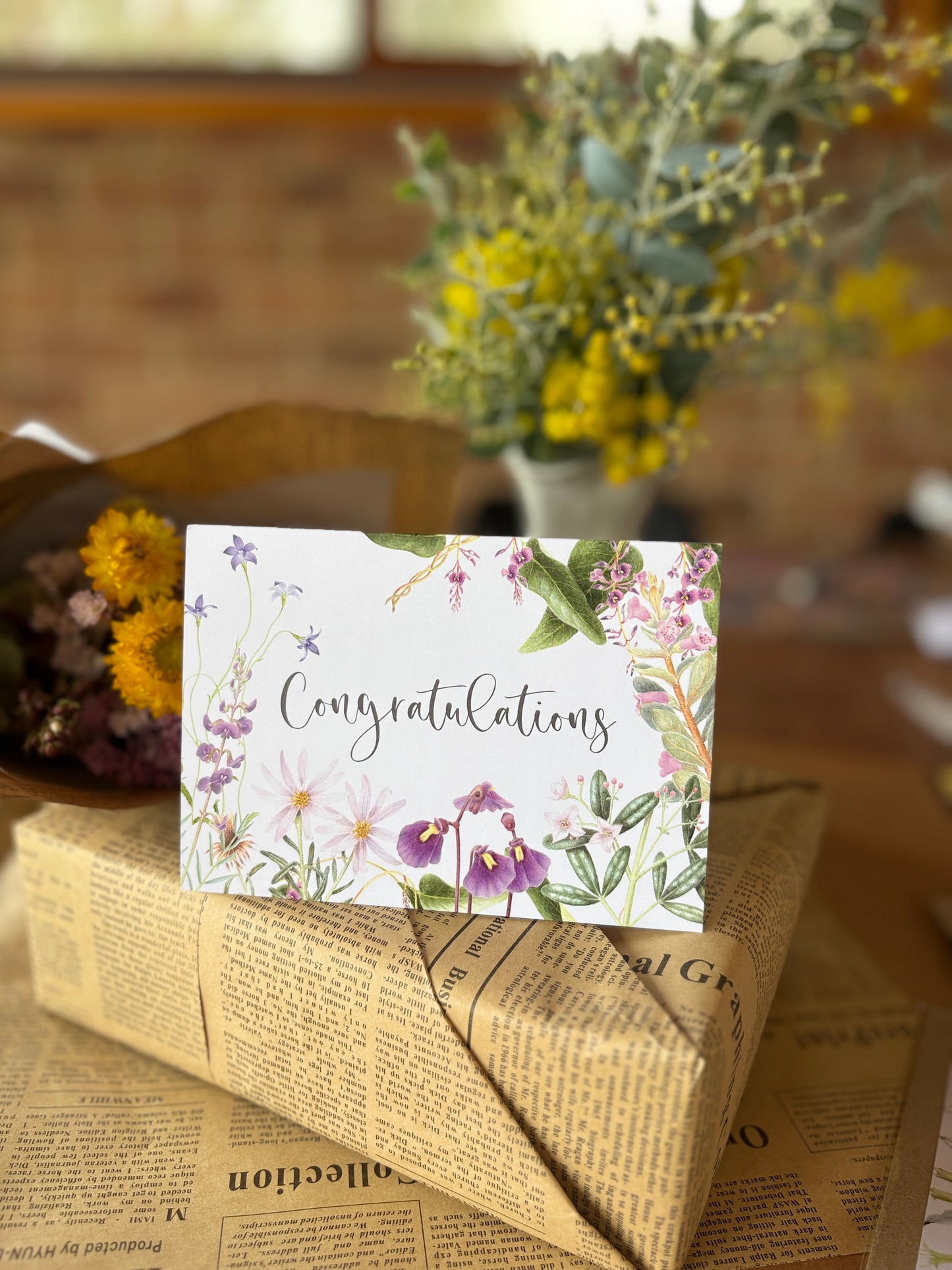 A floral congratulations card with the word 'Congratulations' in script, placed on top of a wrapped package wrapped in newspaper-style paper, with blurred flowers in the background.