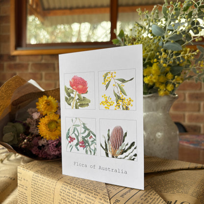 A white greeting card featuring four botanical illustrations of Australian flora, including a pink warbler flower, yellow wattle blossoms, green eucalyptus, and a brown bank flower. The card is printed 'Flora of Australia' and styled on a newspaper with dried flowers in the background.