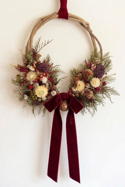 A rustic dried flower wreath hanging on a beige wall, featuring natural driftwood branches, green pine branches, burgundy berries, cream daisies, and olive-green ribbon accents.
