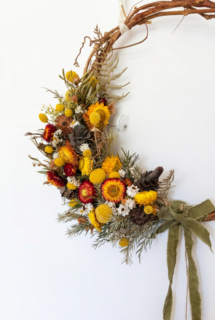 A rustic dried floral wreath made from a grapevine frame, decorated with yellow and orange flowers, green foliage, pinecones, and a green ribbon bow, hanging on a plain off-white wall.