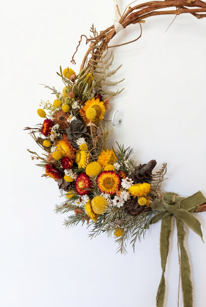 A rustic dried floral wreath made from a grapevine frame, decorated with yellow and orange flowers, green foliage, pinecones, and a green ribbon bow, hanging on a plain off-white wall.
