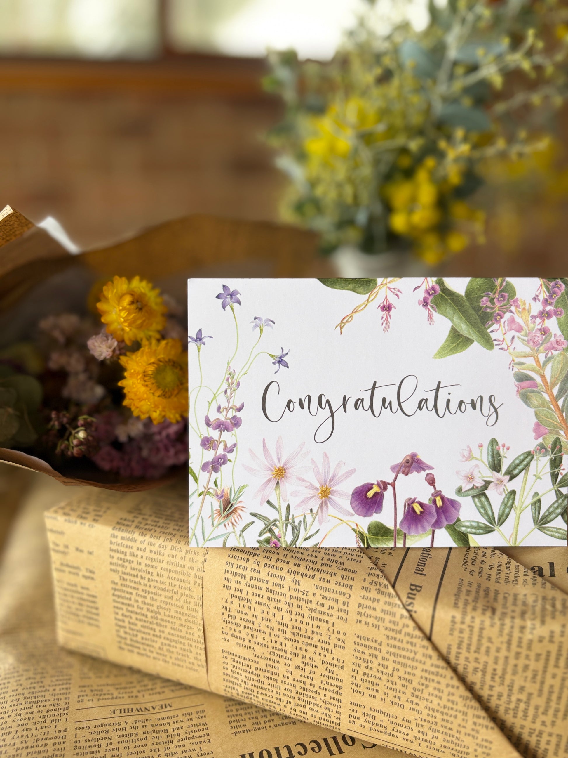 A floral congratulations card with the word 'Congratulations' in script, placed on top of newspaper-wrapped packages with dried flowers in the background.