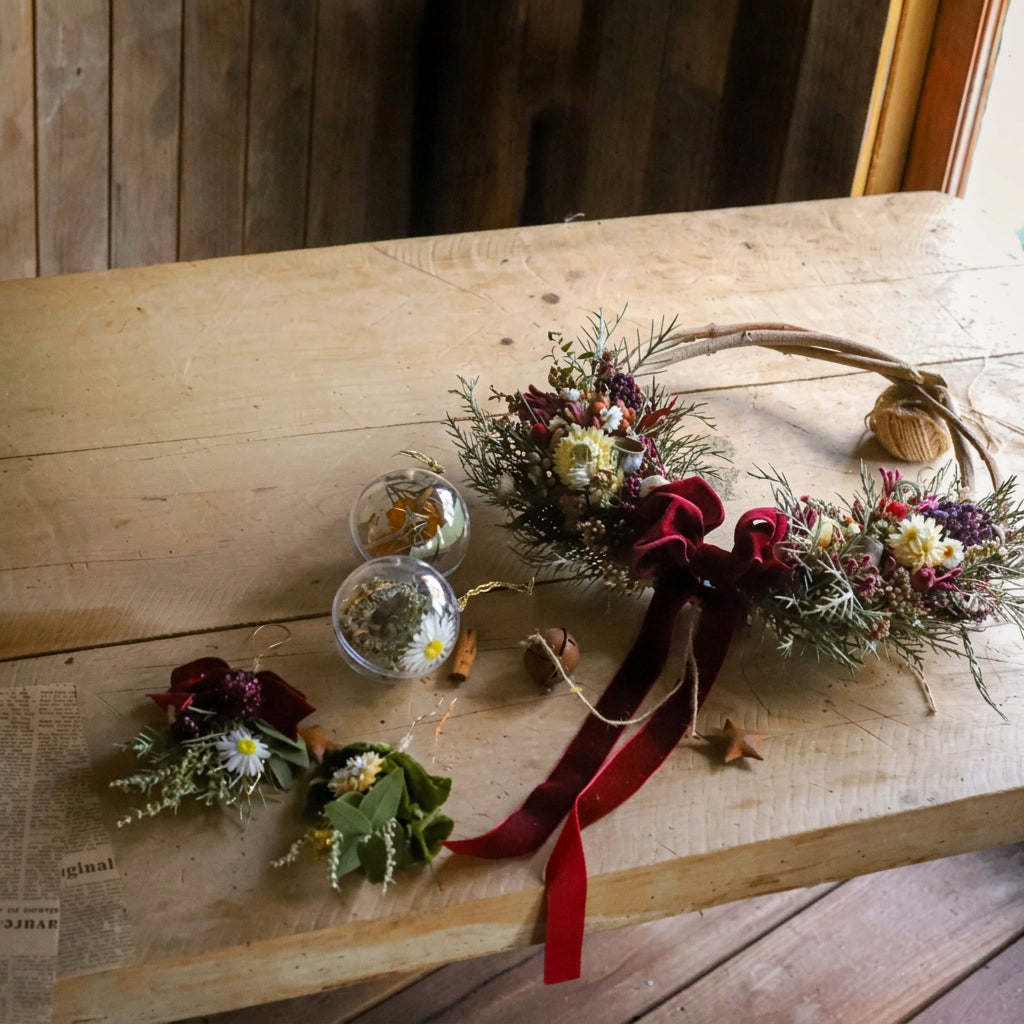 A rustic dried flower wreath with a long red velvet ribbon bow, displayed on a light wooden table alongside two small glass globes and a small floral sprig.