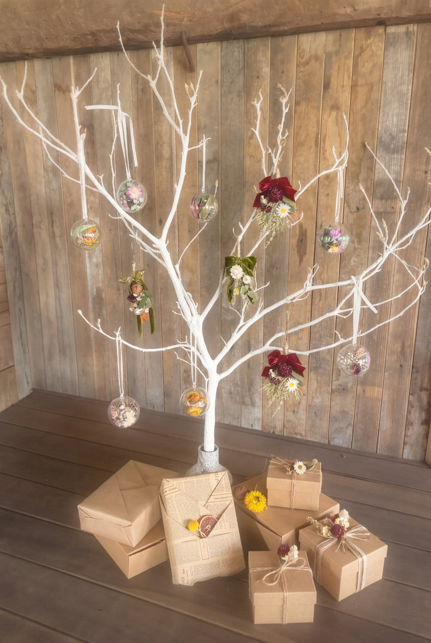 A decorative bare tree stand holding multiple hanging glass ornaments and small floral accents, styled on a dark wooden surface with wrapped gift boxes in front, against a rustic wood plank wall.