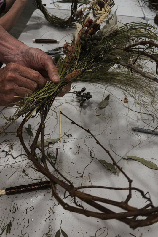 Person crafting a wreath with natural materials on a table.