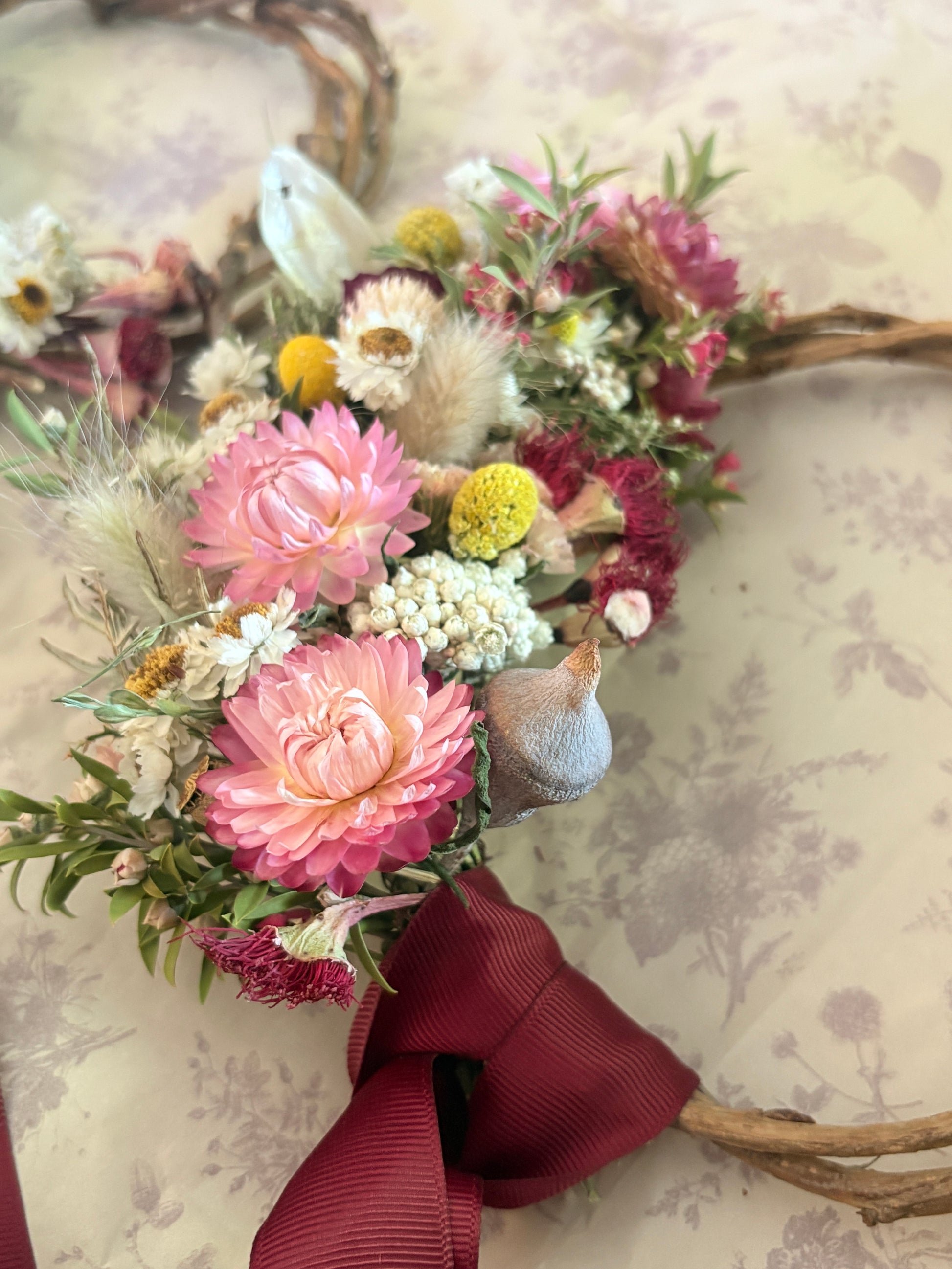 Floral wreath with pink flowers, greenery, and a red ribbon on a textured wall background.