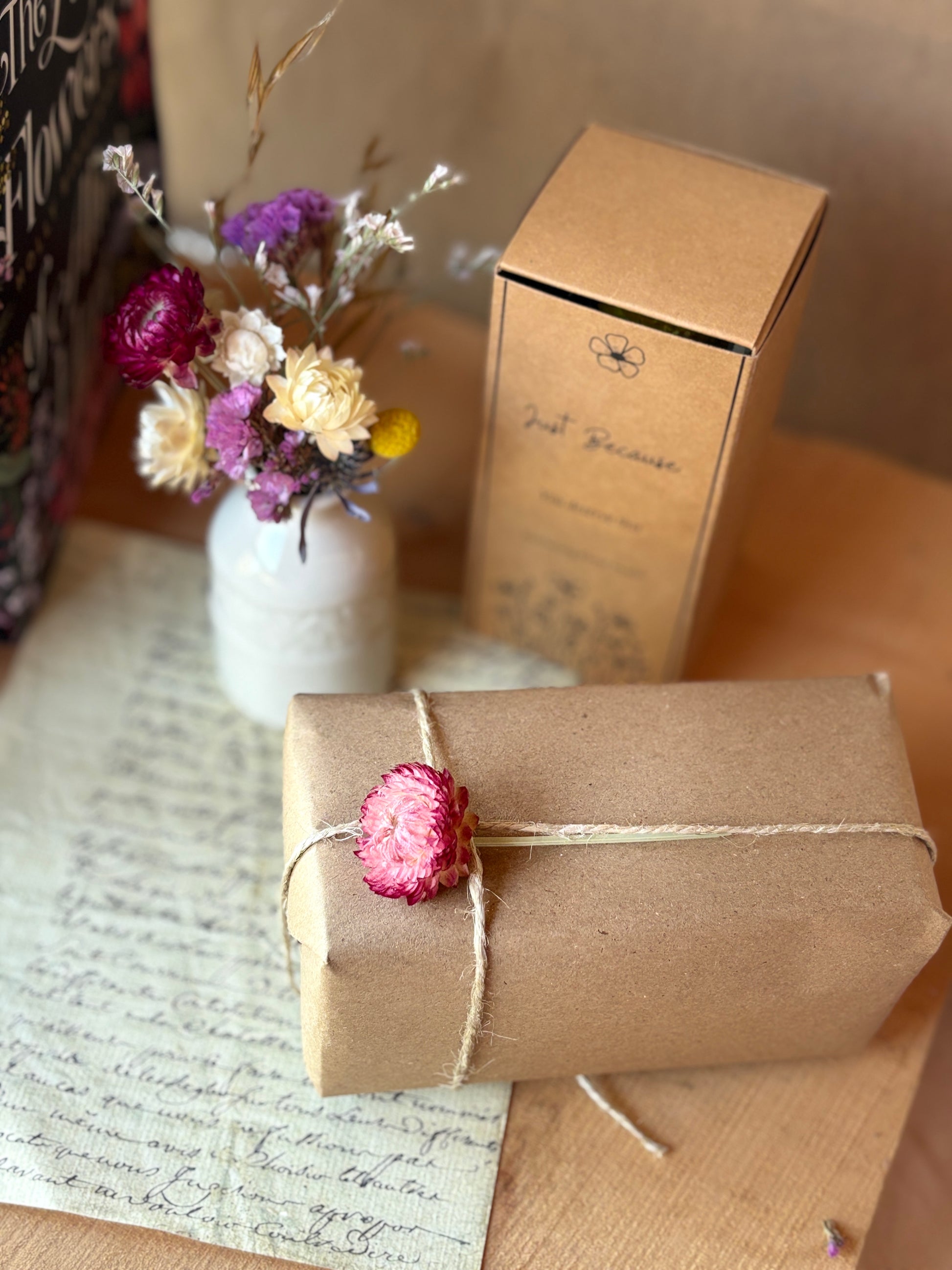 Two brown paper-wrapped packages with floral decorations on a surface with a vase of flowers in the background.