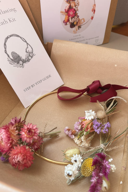 A dried flower crown kit displayed inside its kraft paper packaging, featuring a gold metal headband decorated with pink, purple, white, and yellow dried flowers, a dark red ribbon, a step-by-step guide card, and a floral instruction booklet.